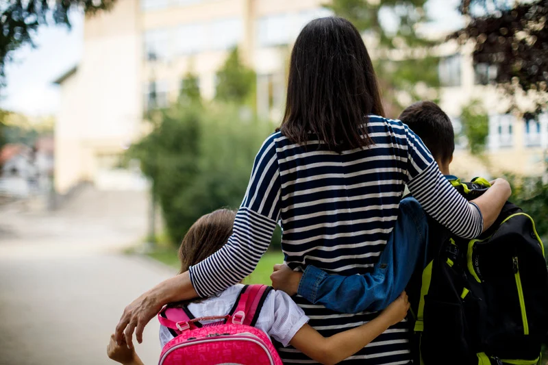 Madre caminando con sus hijos hacia el colegio, abrazándolos por los hombros.