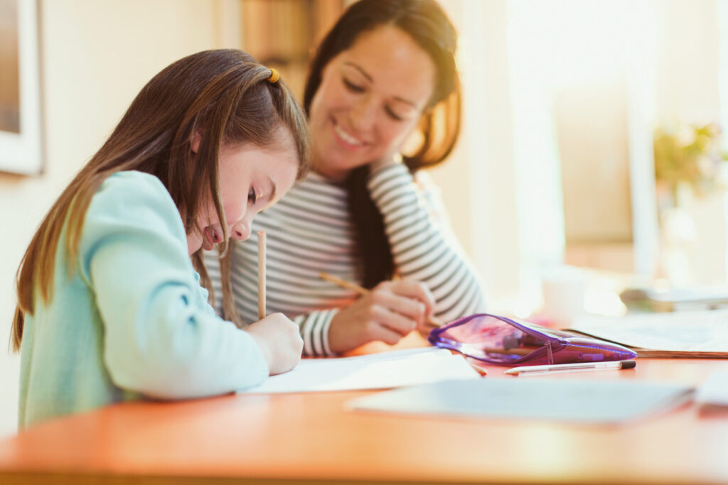 Madre apoyando a su hija mientras escribe en sus tareas escolares, representando un inicio de estudio tranquilo y motivado.