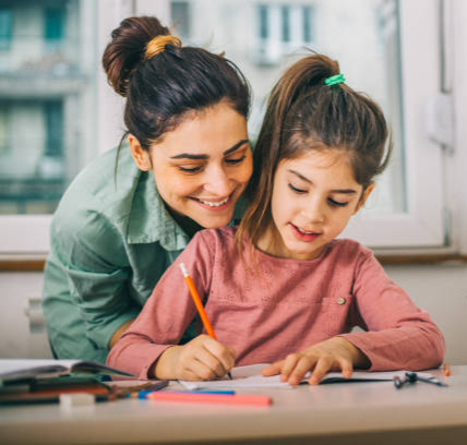 Una nina está haciendo los deberes contenta. Su madre mira por encima de su hombro sonriendo simbolizando que los deberes pueden ser un momento de conexión.