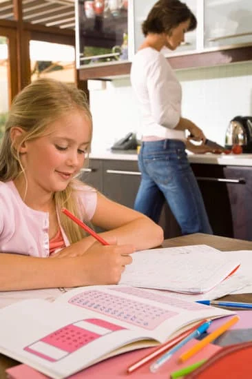 Niña haciendo los deberes de forma autónoma mientras su madre cocina cerca, accesible pero sin intervenir